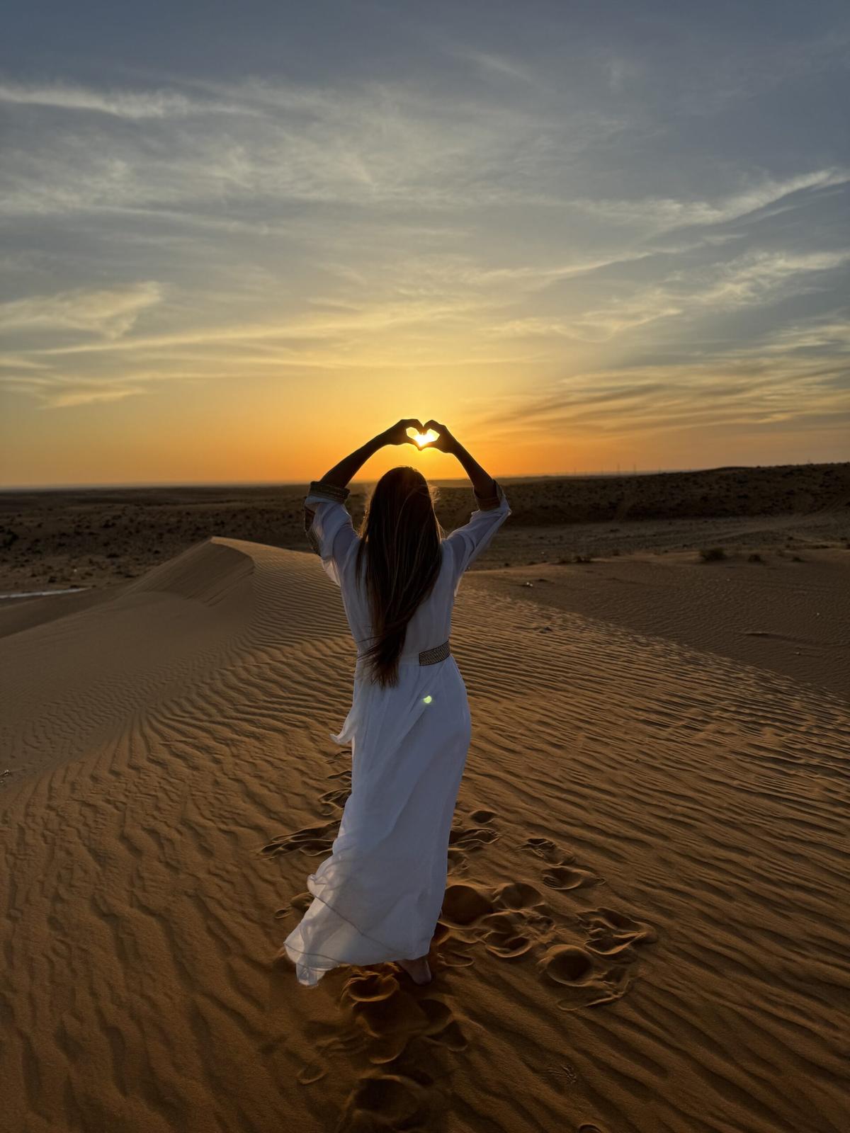 Woman in white dress making heart shape with hands at sunset in desert